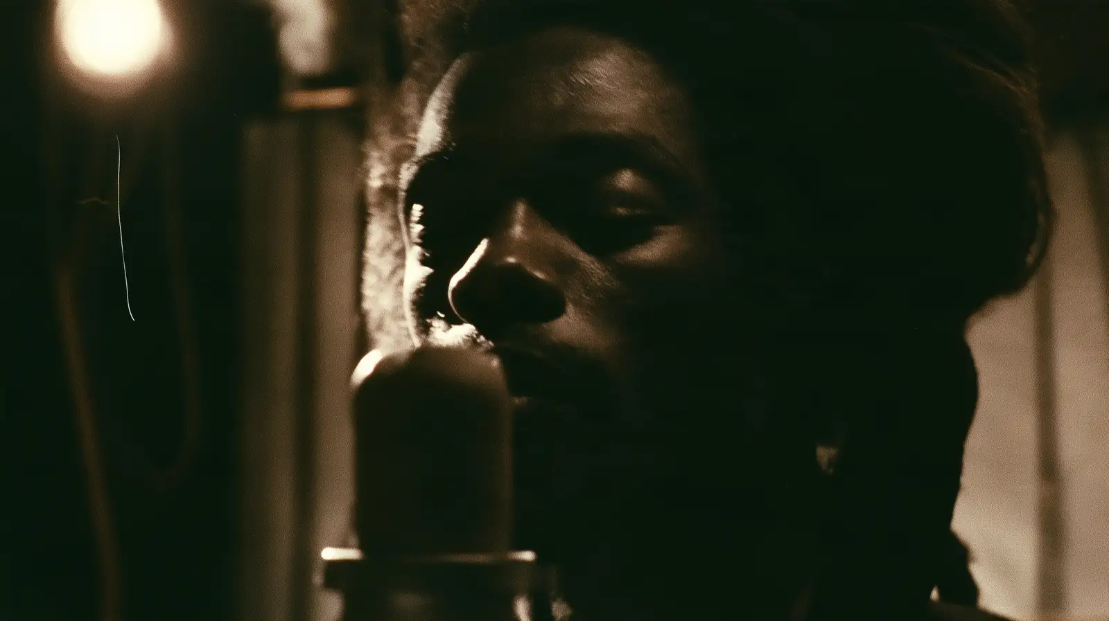 A singer close to a large studio microphone in a darkened room, lit by one warm lamp, caught mid phrase with eyes lowered.
