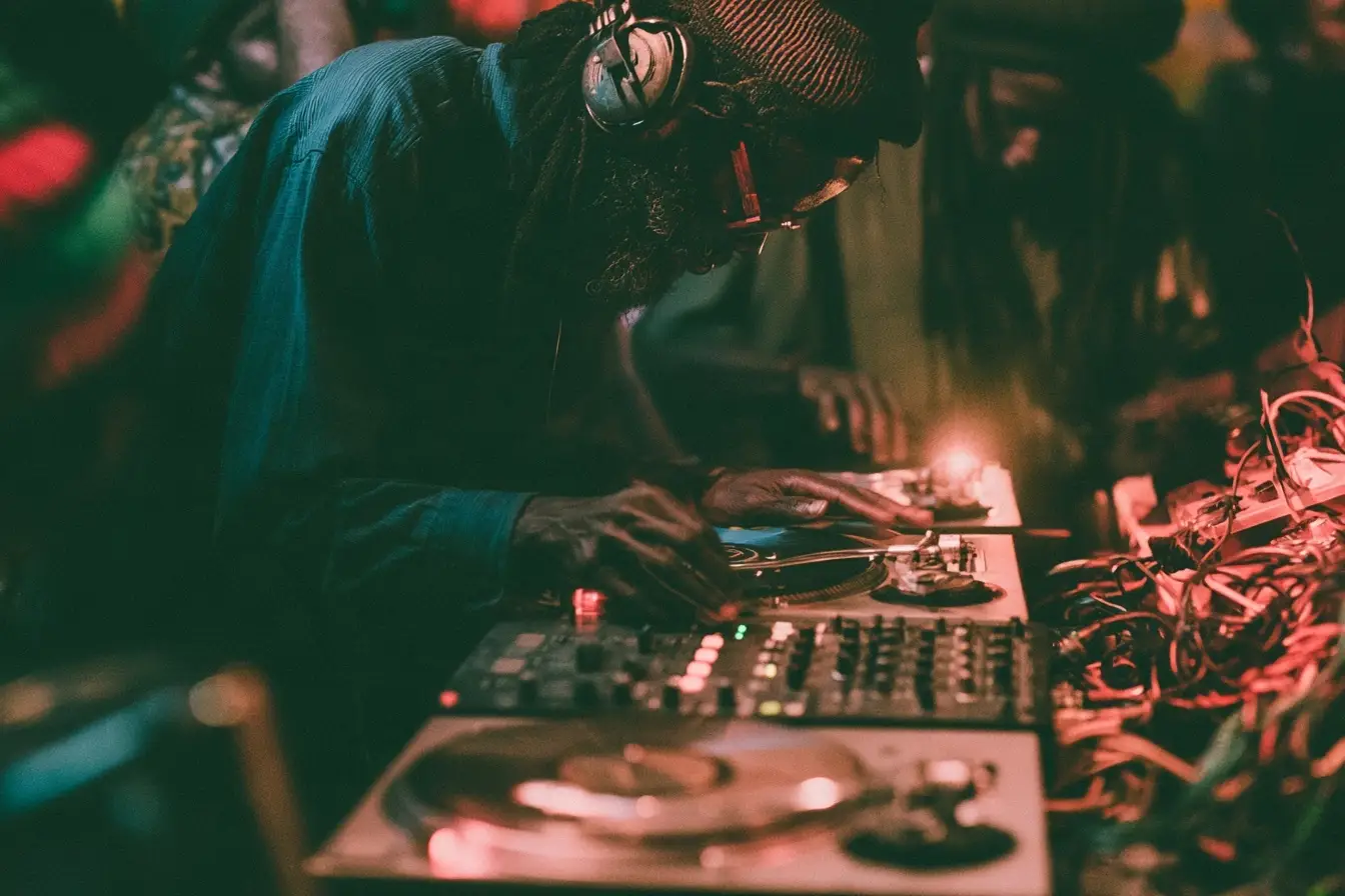 A selector at the controls in a dark hall, lit red and green, hands on the mixer beside a turntable and a tangle of cable.